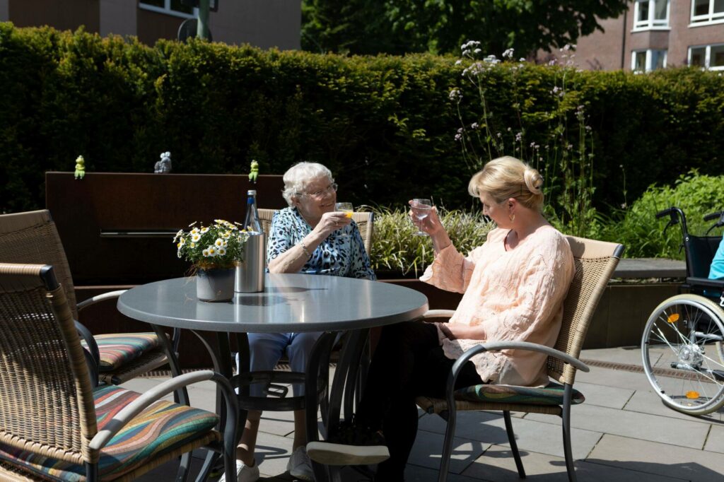 Auf der Sonnenterrasse der stationären Pflege: Eine Seniorin prostet einer jungen Frau zu.