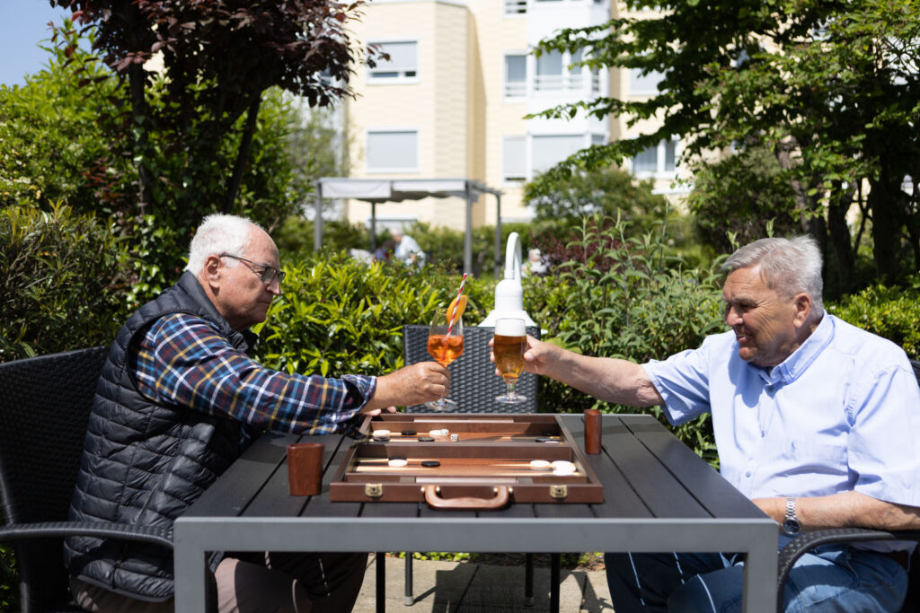 Zwei Herren sitzen auf der sonnigen Dachterrass, spielen Backgammon und prosten sich mit Bier bzw. einem Aperol zu.